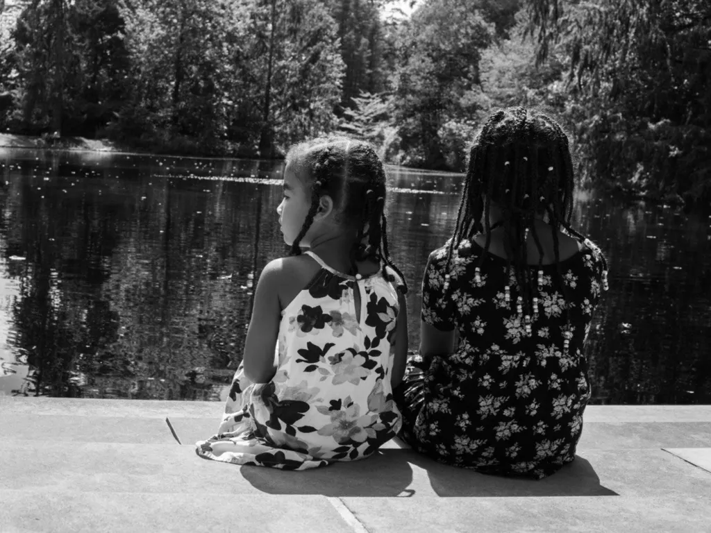 Black and white photo of the two sisters from behind, sitting side by side at the edge of a pond in matching floral dresses